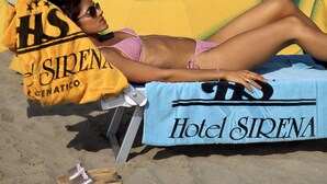 A person lying on a beach chair under a large umbrella with the text "Hotel Sirena" on a towel. - Hotel Sirena (Cesenatico)