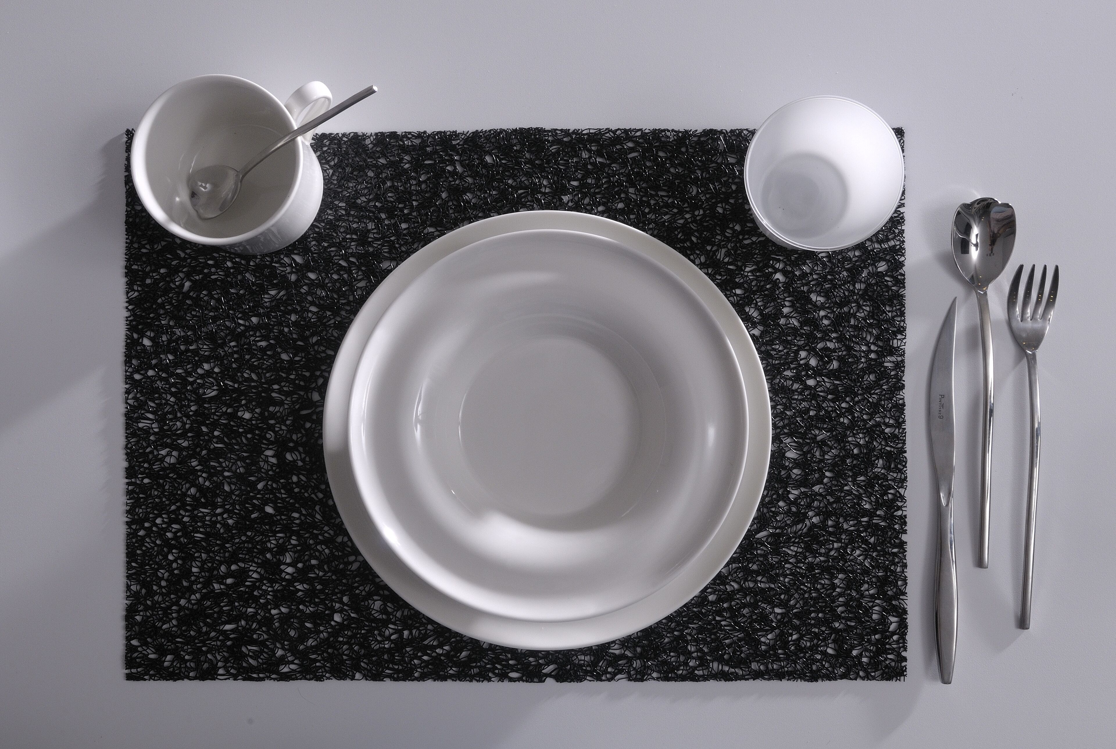Une table dressée avec une assiette blanche, une tasse blanche, une cuillère, une fourchette et un couteau, le tout posé sur une nappe noire à texture.