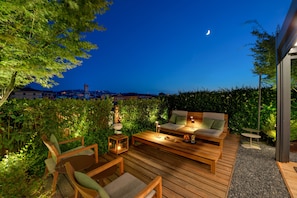 Un patio al aire libre con mobiliario de madera, un sofá y una mesa, rodeado de follaje y un paisaje urbano al fondo.