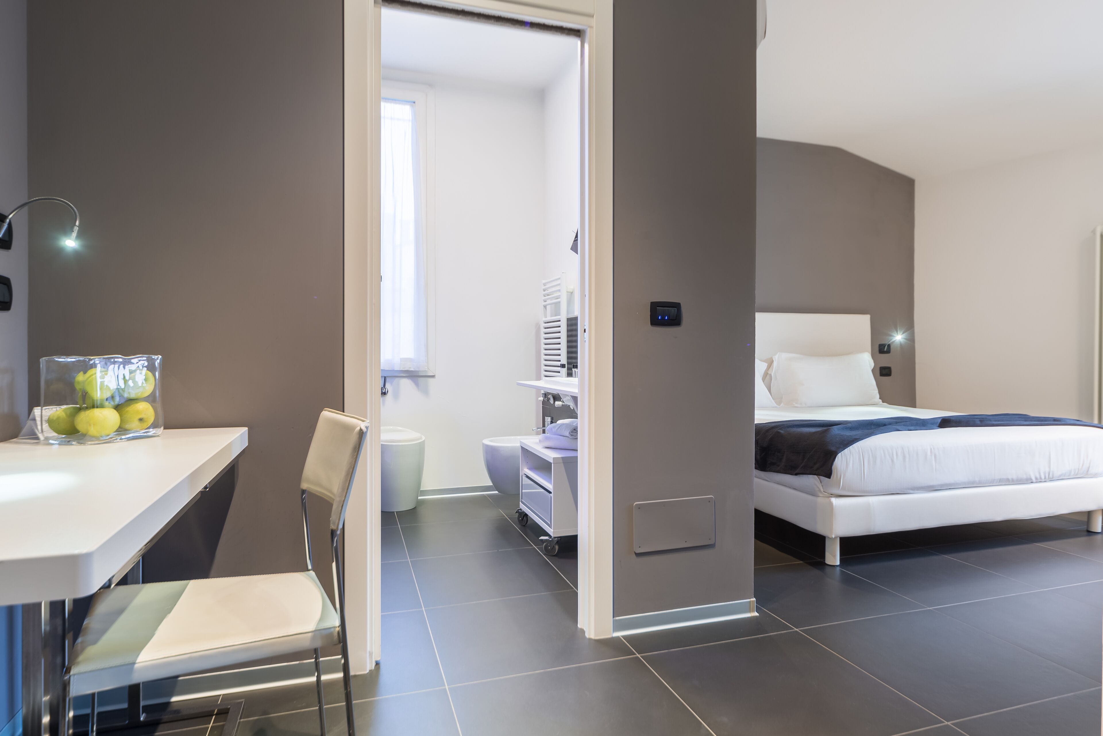 Una camera d'albergo moderna con un letto, una scrivania con una sedia, un tavolino con una ciotola di frutta e un bagno visibile attraverso una porta aperta.