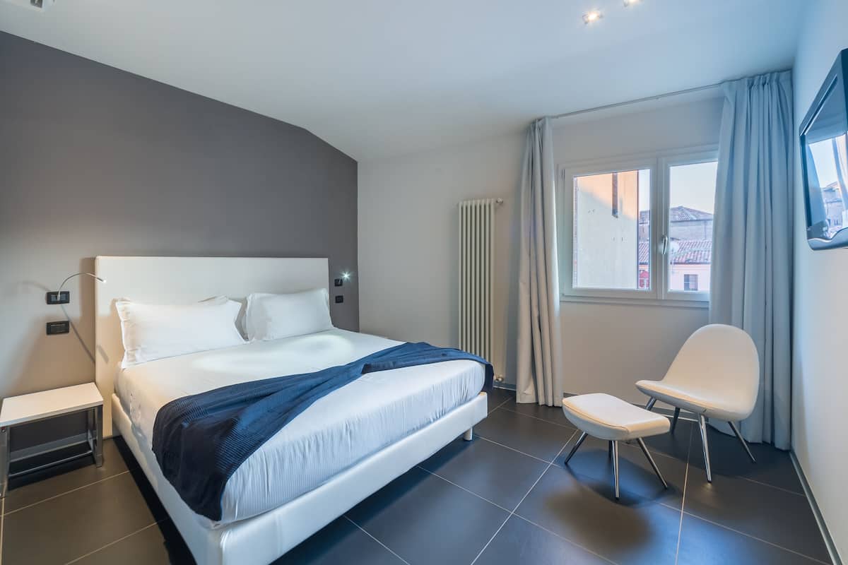 Ein modernes Hotelzimmer mit einem großen Bett, einem Nachttisch, einem Sessel und einem Fenster mit Vorhängen.