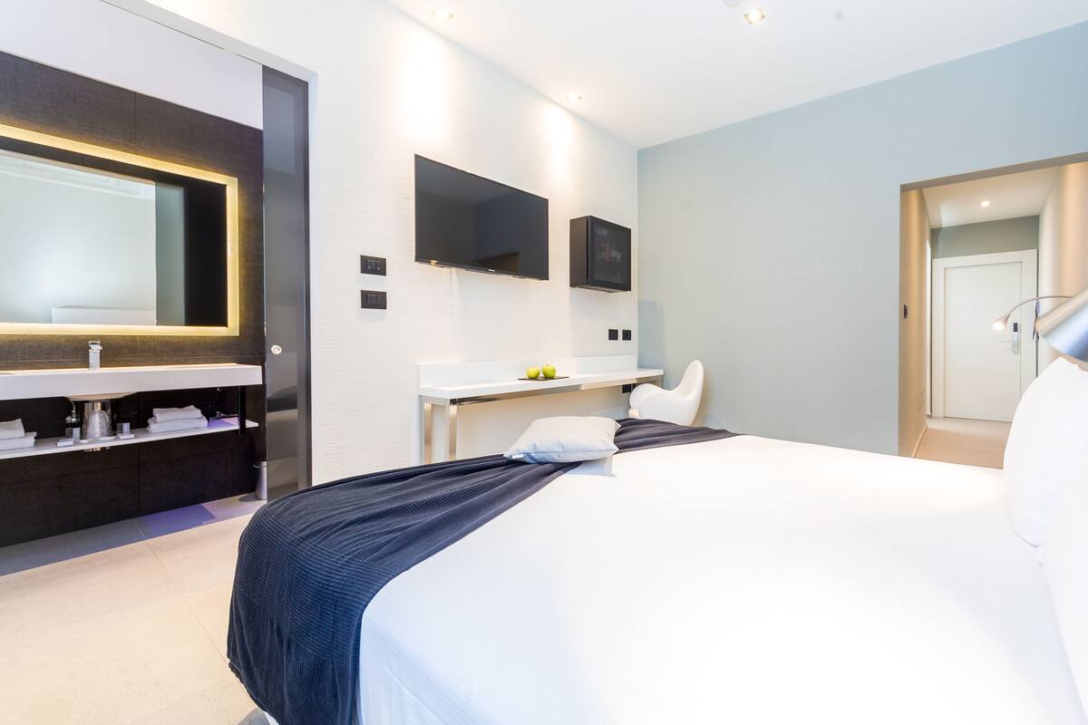 Ein modernes Hotelzimmer mit einem großen Bett, einem Nachttisch, einem Schreibtisch mit Stuhl, einem großen Spiegel und einem Fernseher.