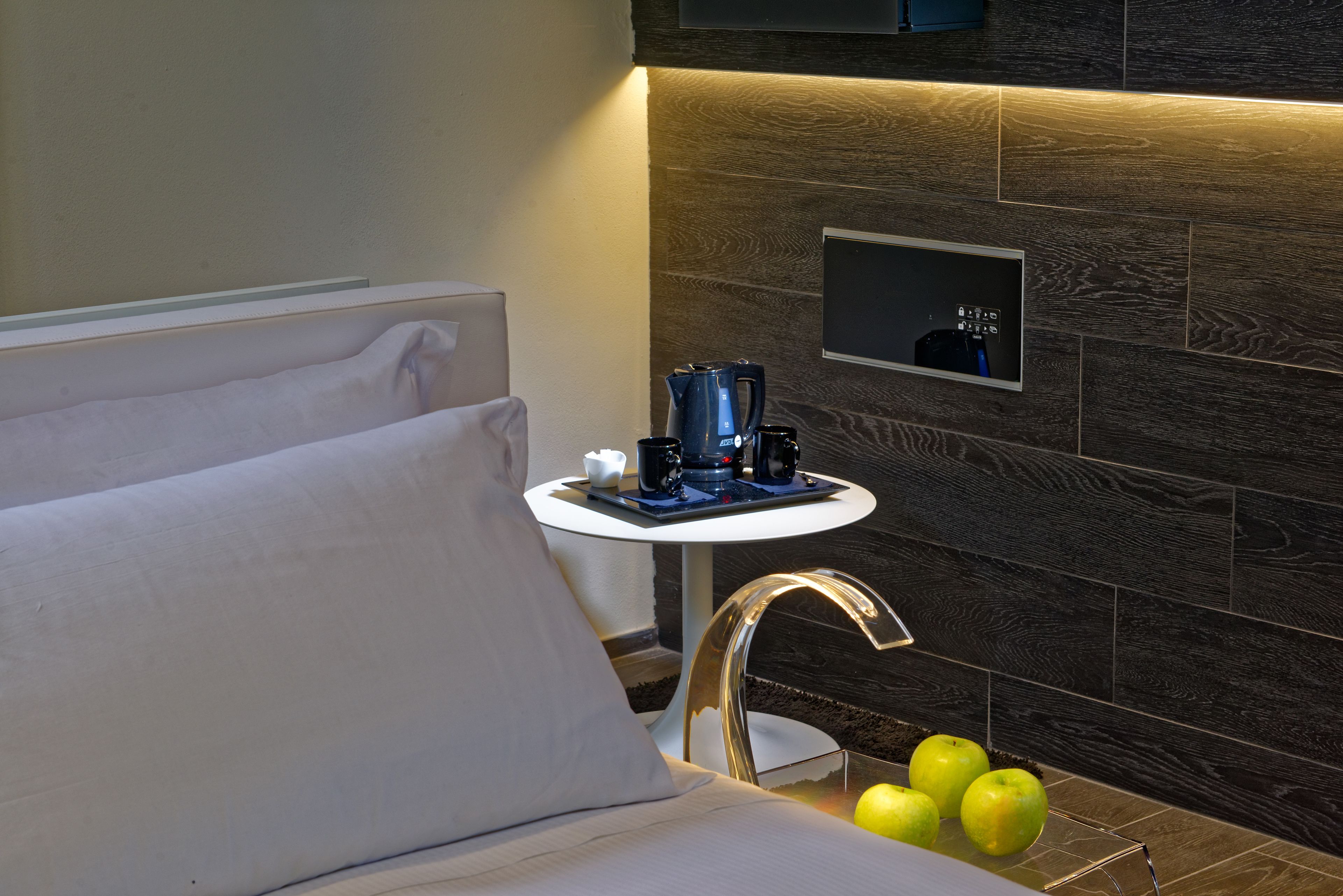 A modern hotel room with a bed, a small table with a coffee maker, and a wall-mounted control panel.