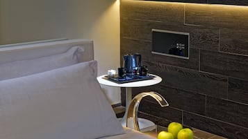 A modern hotel room with a bed, a small table with a coffee maker, and a wall-mounted control panel.