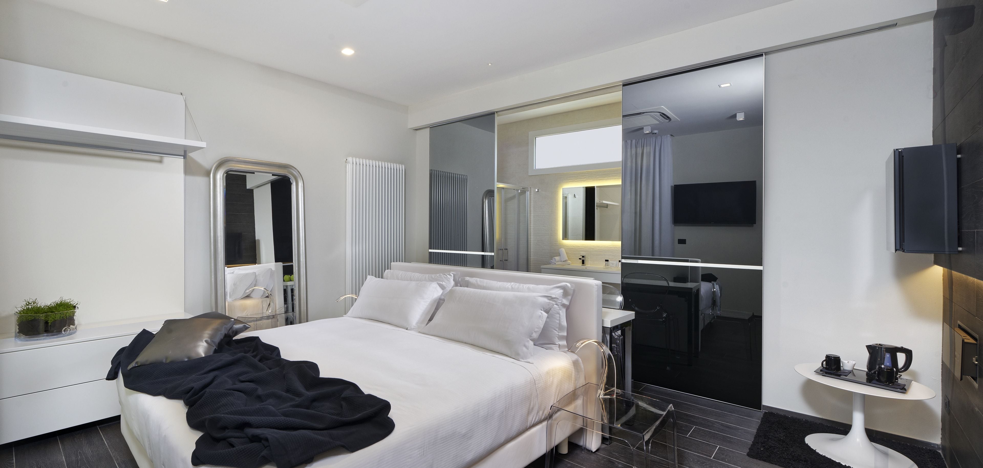 Een moderne hotelkamer met een groot bed, een nachtkastje met een plant, een spiegel, een televisie en een klein tafeltje met een koffiezetapparaat.