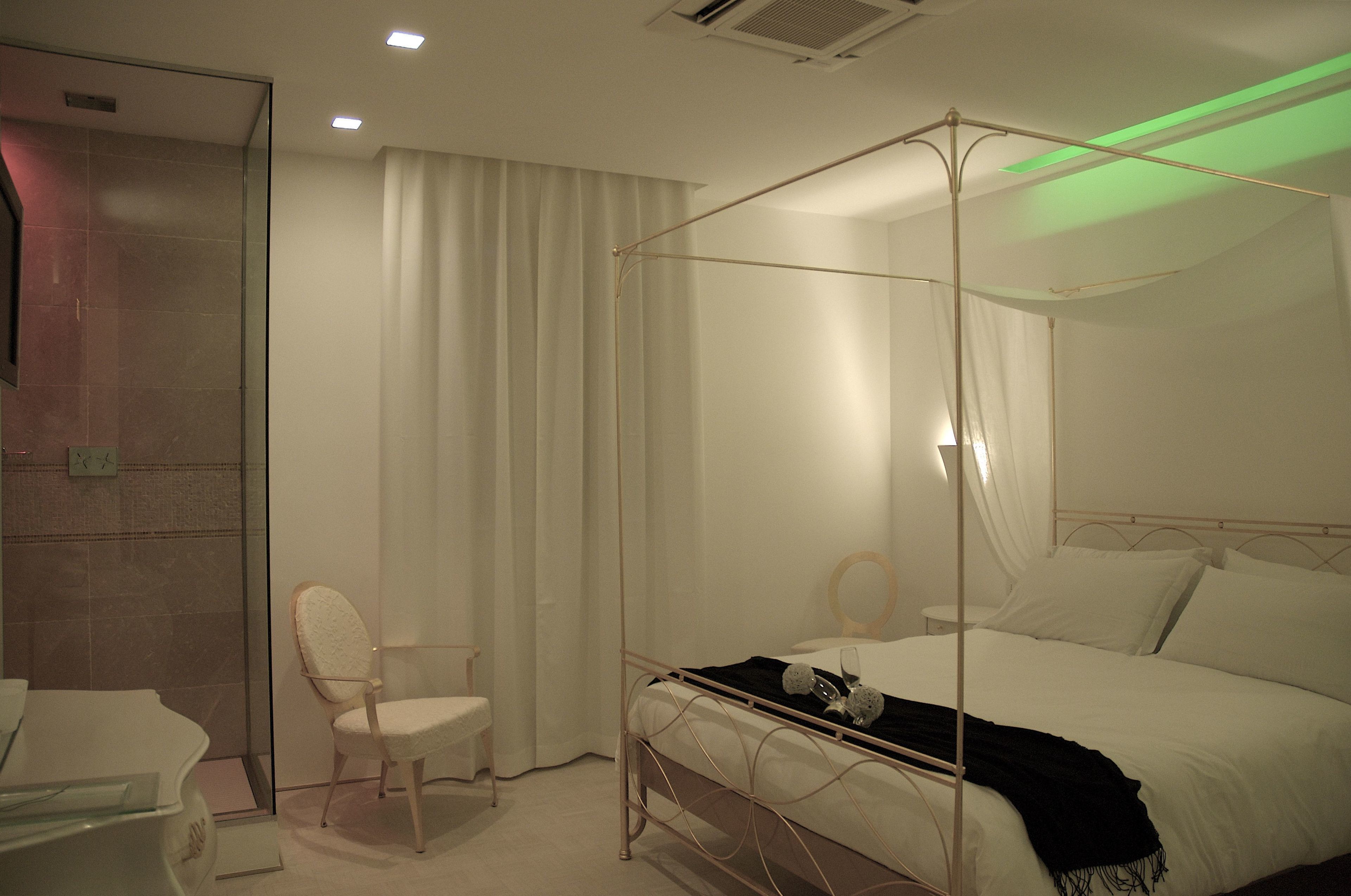 A four-poster bed with white linens, a bedside table, a chair, and a glass shower enclosure.