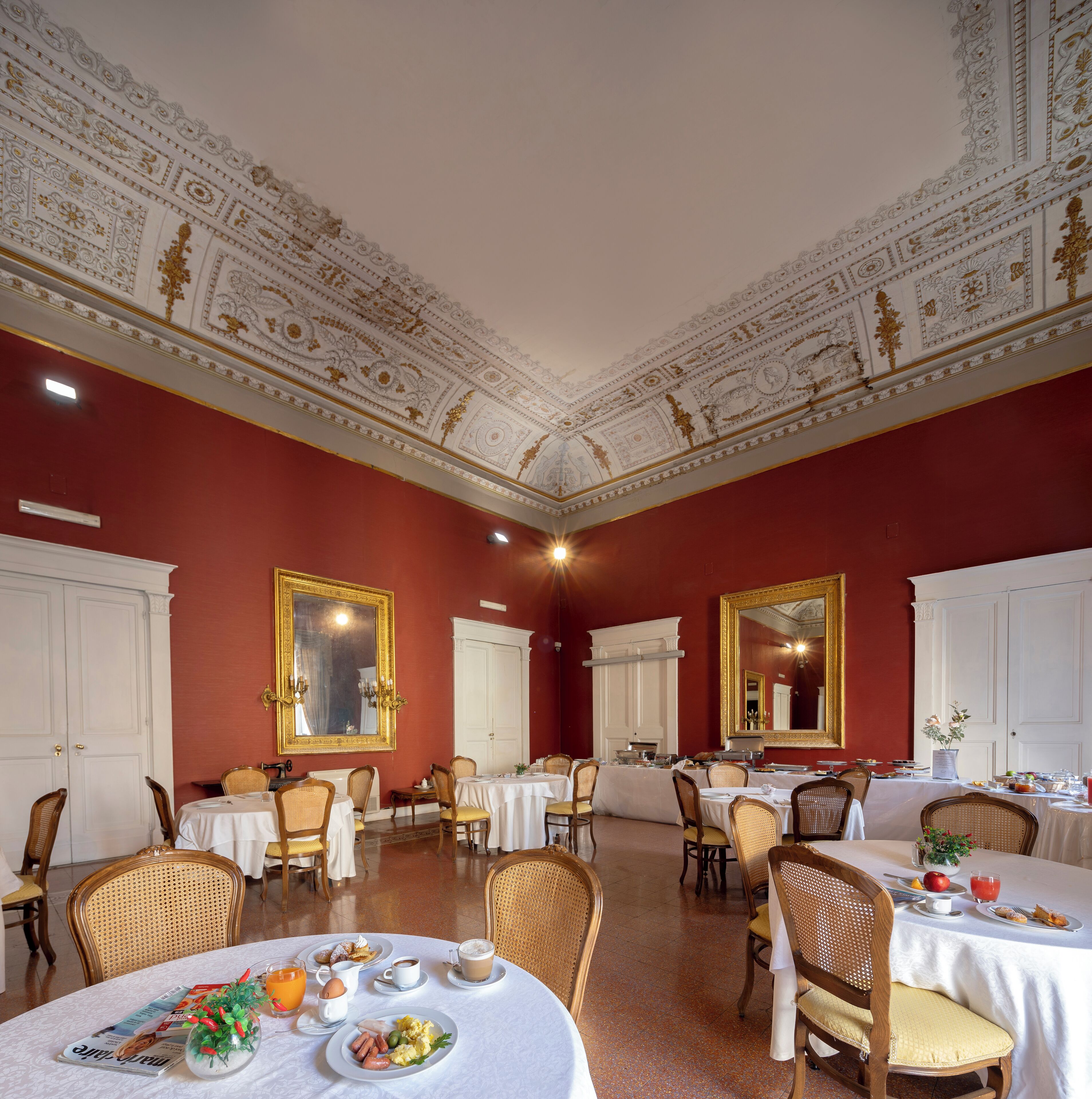 Une salle à manger agrémentée de décorations élaborées au plafond, de murs blancs et de touches de rouge. Les tables sont dressées avec des aliments pour le petit-déjeuner, et plusieurs chaises sont disposées autour de chaque table.