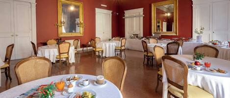 Une salle à manger agrémentée de décorations élaborées au plafond, de murs blancs et de touches de rouge. Les tables sont dressées avec des aliments pour le petit-déjeuner, et plusieurs chaises sont disposées autour de chaque table.