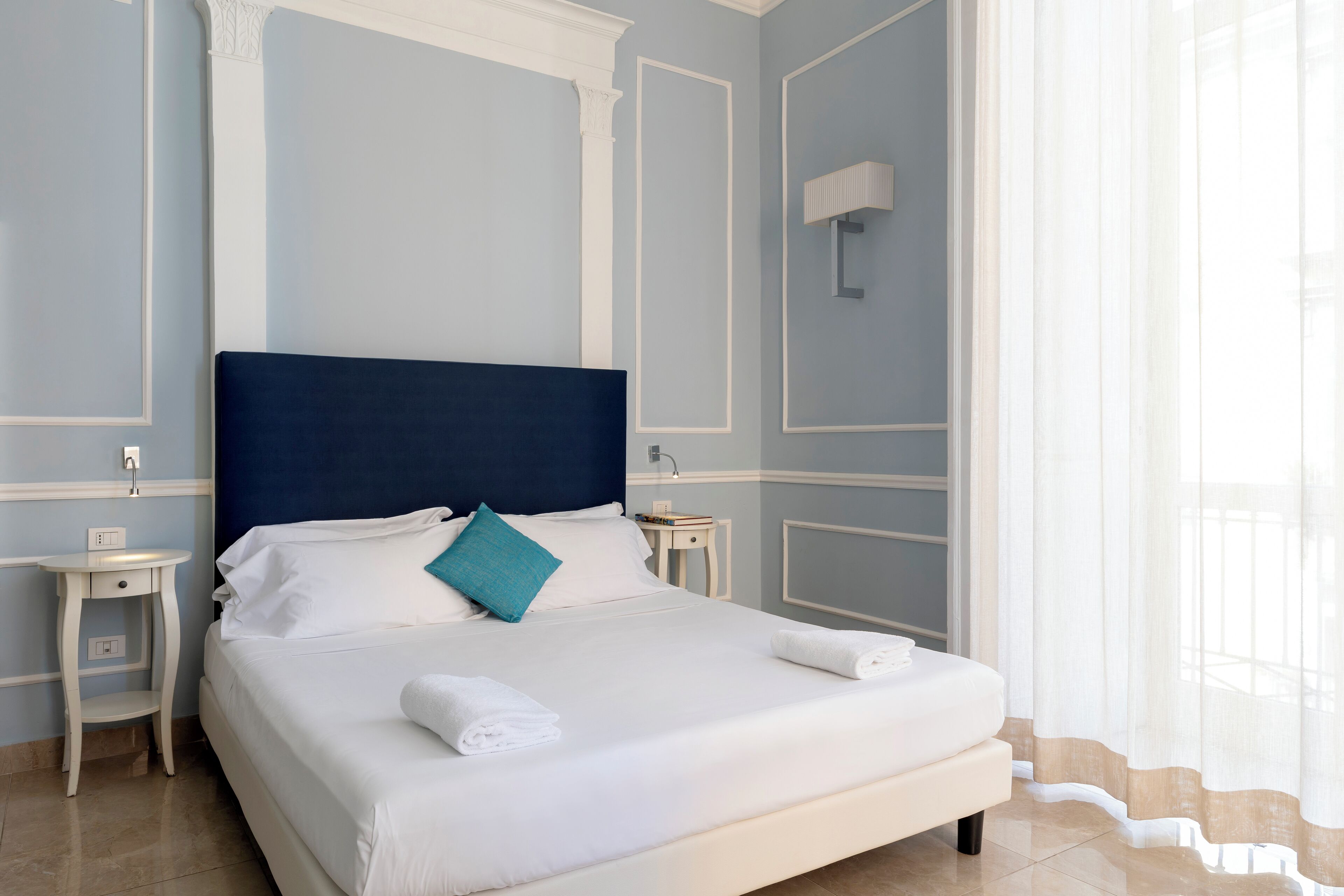 A neatly made bed with white linens and a blue headboard, two bedside tables, and a window with sheer curtains.