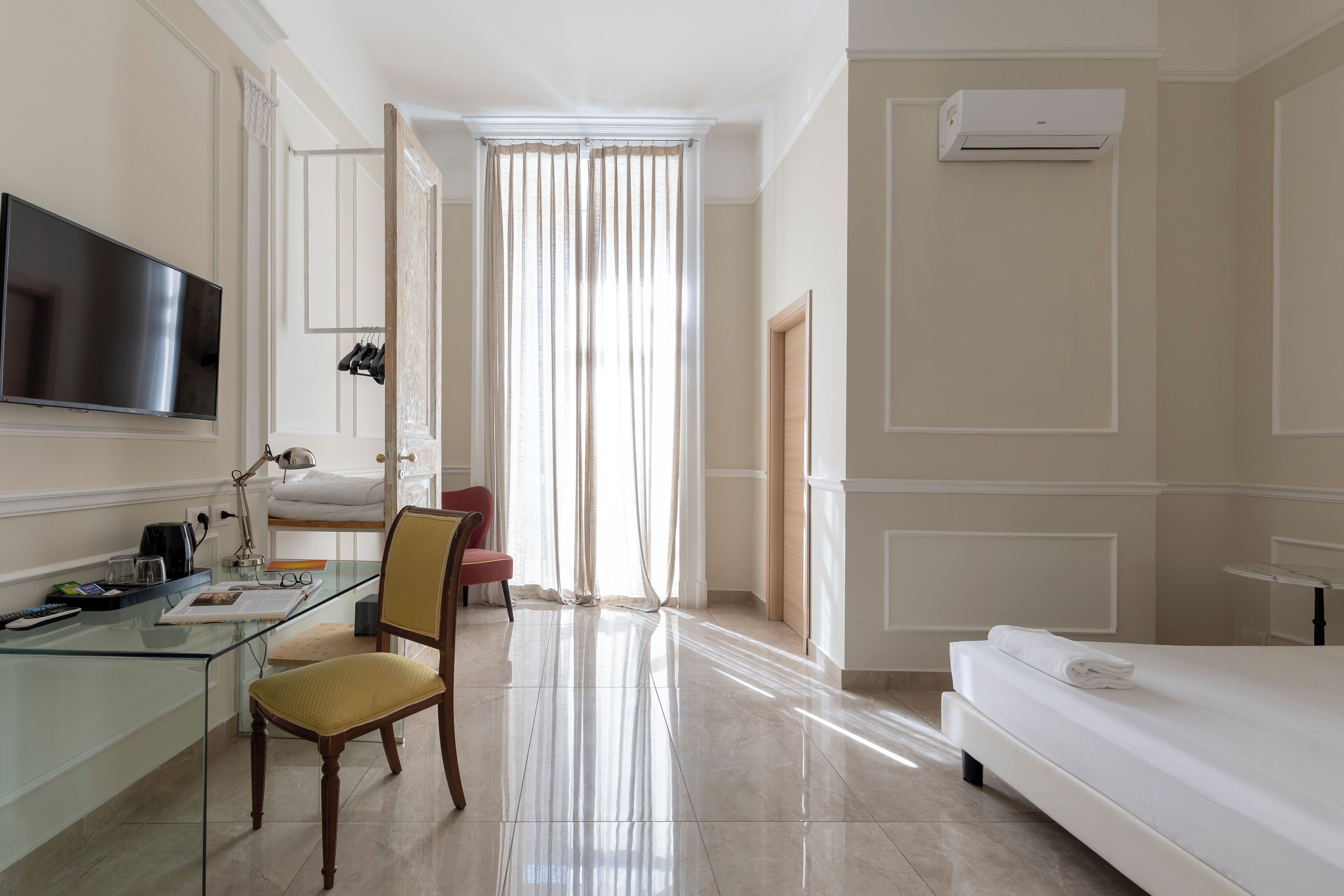 A hotel room with a glass desk, a chair, a bed, a TV, and an air conditioning unit.