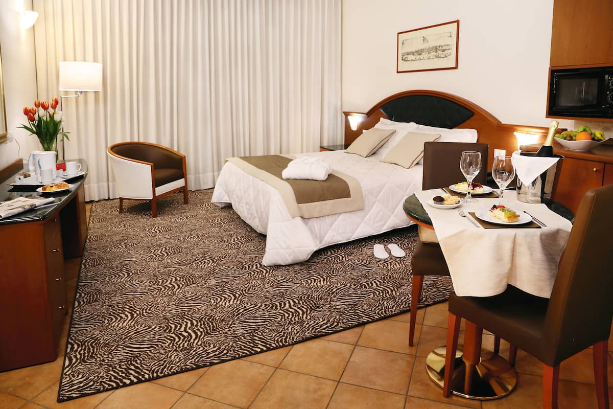 A hotel room with a bed, a desk with a chair, a dining table with a chair, a television, and a window with curtains.