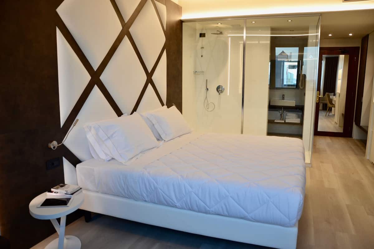 A modern bedroom with a large bed, a bedside table with a book, and a glass-enclosed shower area.