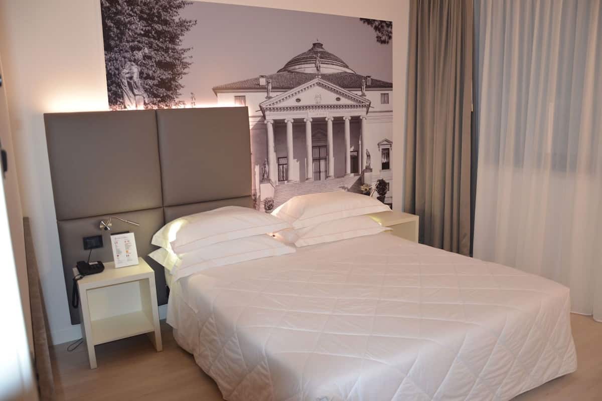 A hotel room with a large bed, a nightstand with a lamp, a wall-mounted TV, and a poster of a classical building.