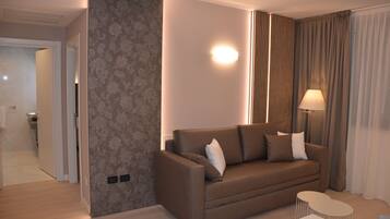 A modern living room with a brown sofa, a small white table, and a lamp. There is a door leading to another room and a wall with a floral pattern.