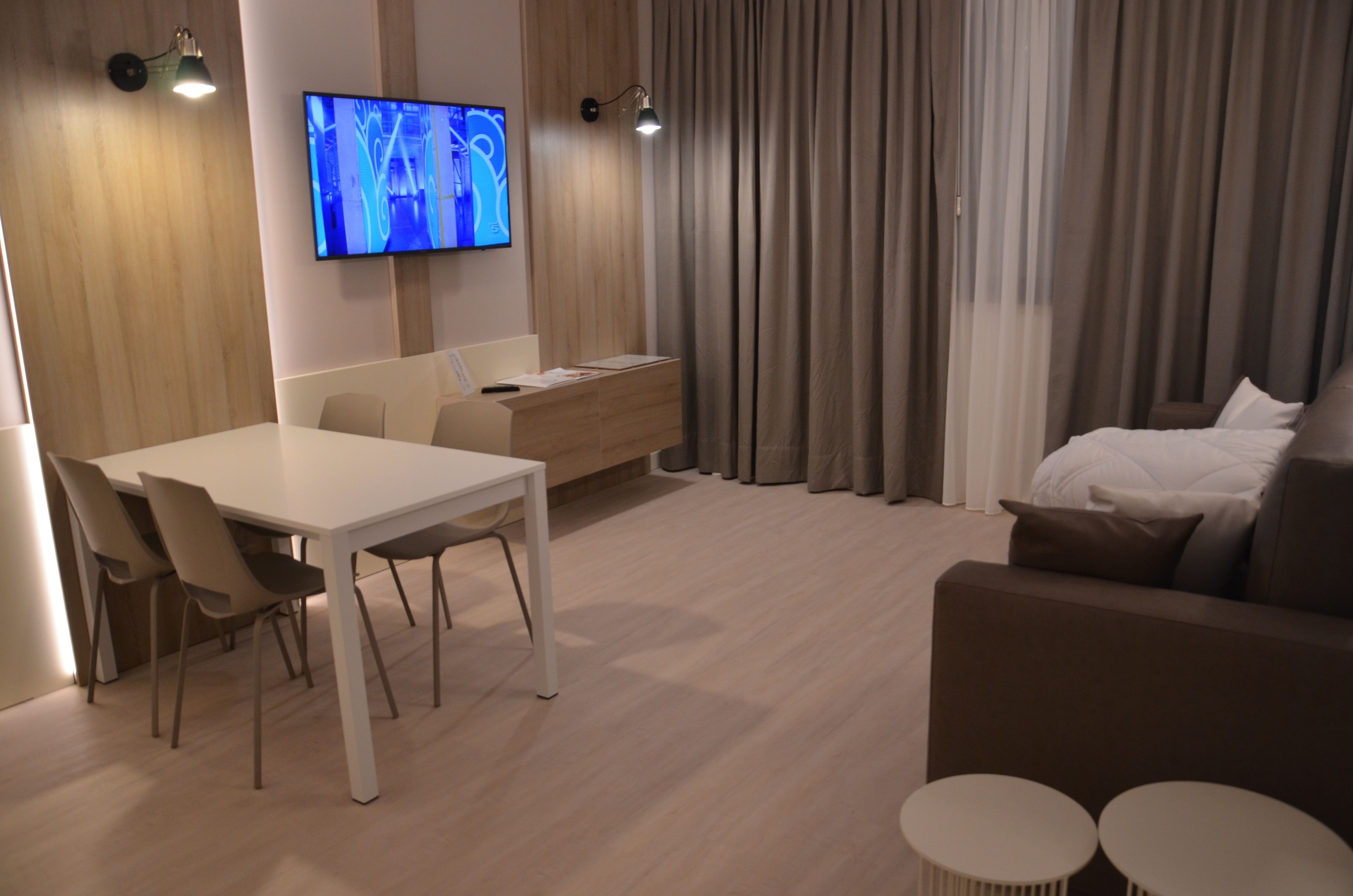 A modern hotel room with a dining area, a sofa, and a TV on the wall.