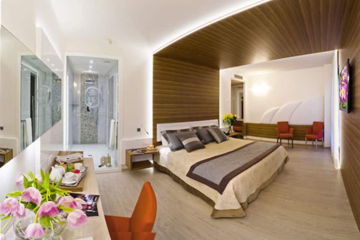 A modern bedroom with a large bed, a sitting area with chairs, and a bathroom visible through a glass door.