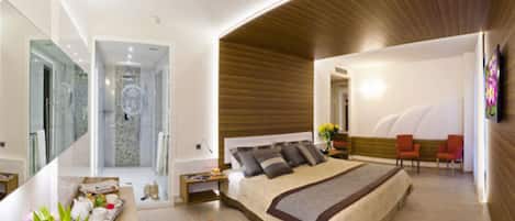 A modern bedroom with a large bed, a sitting area with chairs, and a bathroom visible through a glass door.