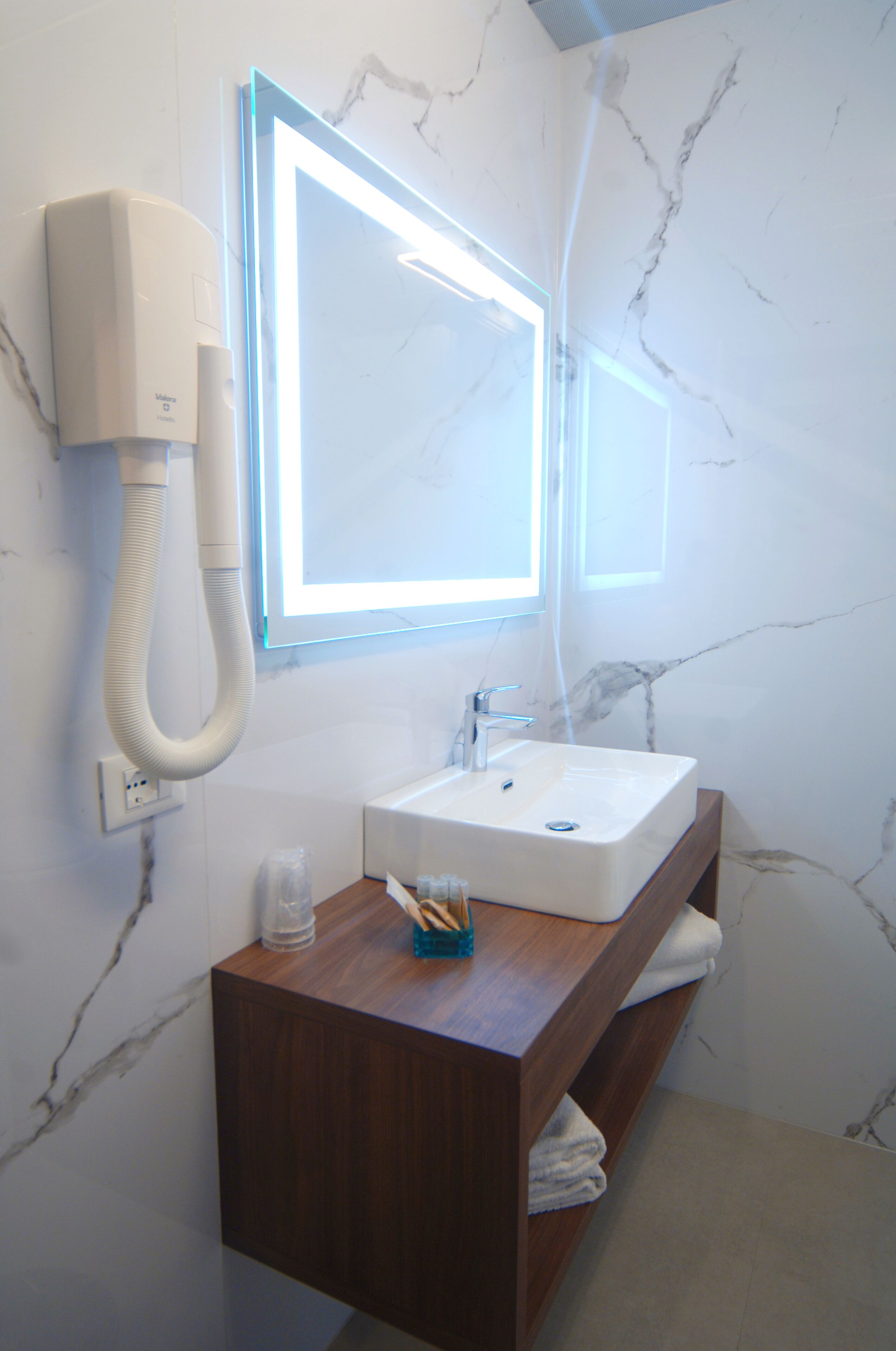 Ein Badezimmer mit Marmorwand, einem weißen Waschbecken, einer Holzkommode, einem Spiegel mit LED-Beleuchtung und einer Duschbrause.