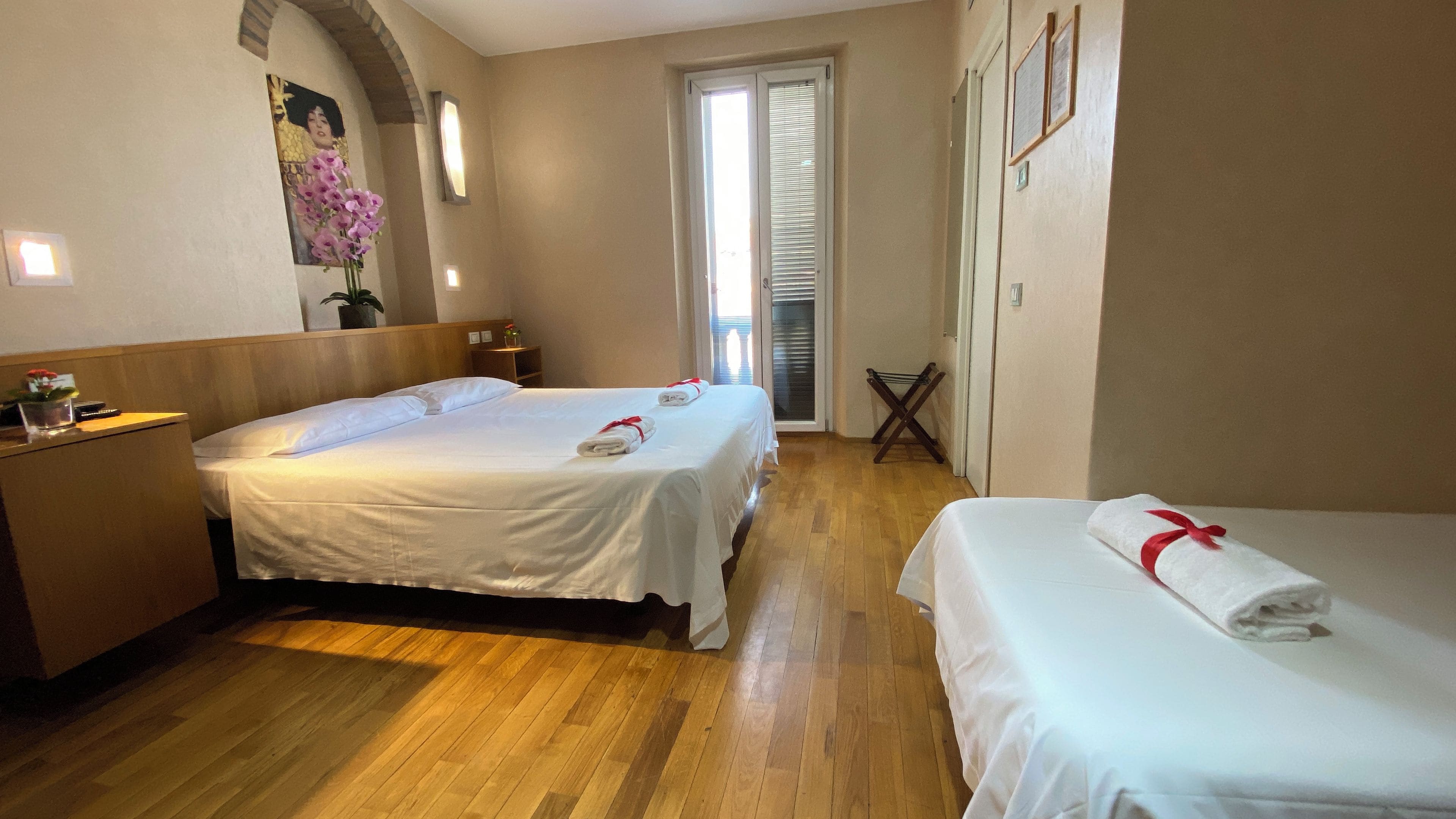 A hotel room with two beds, wooden flooring, a small table with a vase, and a window with blinds.