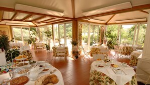 Un amplio comedor al aire libre con varias mesas dispuestas para el desayuno, rodeado de grandes ventanales con vistas a la vegetación.