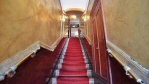 Una imponente escalera con alfombra roja, bordeada por paredes de madera y barandas de metal, que conduce a un ascensor en la parte superior.