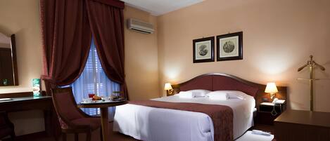 A hotel room with a bed, bedside tables, a desk, a chair, a window with curtains, and two framed pictures on the wall.