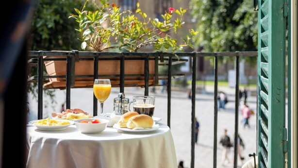 Una mesa dispuesta con comida y bebidas, una planta en maceta, y un edificio histórico al fondo.