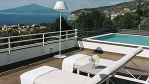 A rooftop terrace with lounge chairs, a pool, and a view of the sea and mountains.