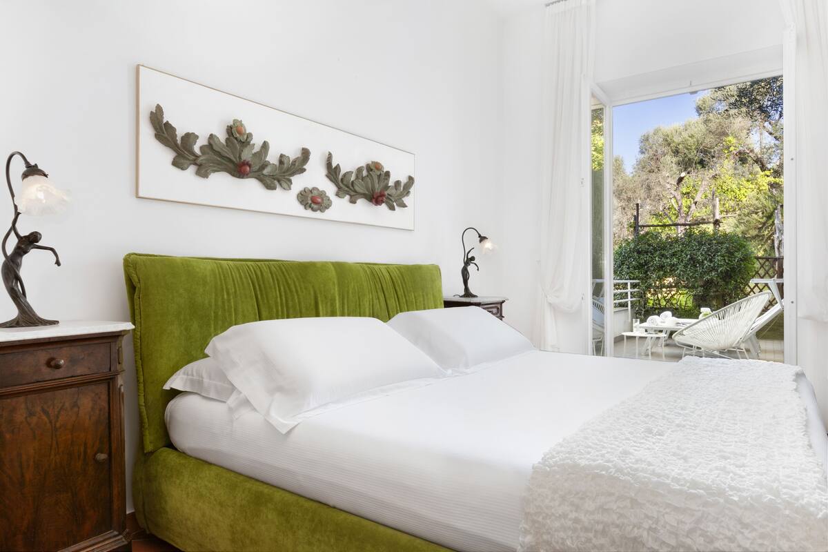 Een slaapkamer met een groen bekleden hoofdeinde, witte beddengoed en een bloemrijk kunstwerk in een lijst aan de muur.