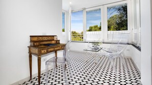 A room with a checkered floor, a wooden side table, a glass table with chairs, and large windows with a view of greenery.