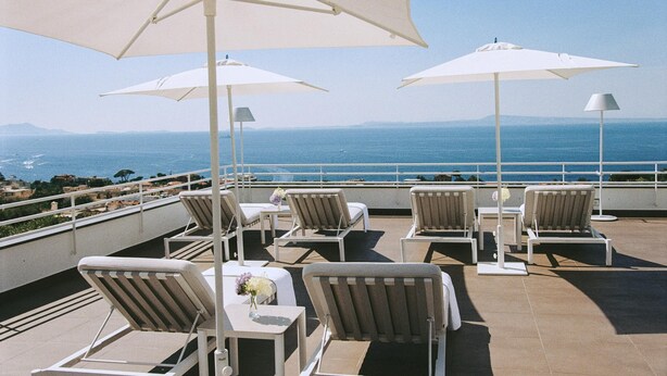 A rooftop terrace with white lounge chairs and umbrellas, overlooking a coastal view.