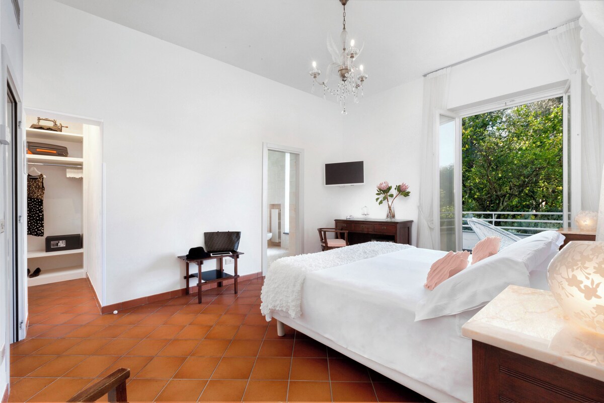 Ein Schlafzimmer mit Bett, Schreibtisch, Stuhl, Fernseher und einem Balkon mit Blick auf Bäume.