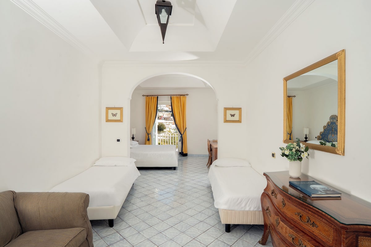 A hotel room with two single beds, a sofa, a dresser, a mirror, and a view of the city through an archway.