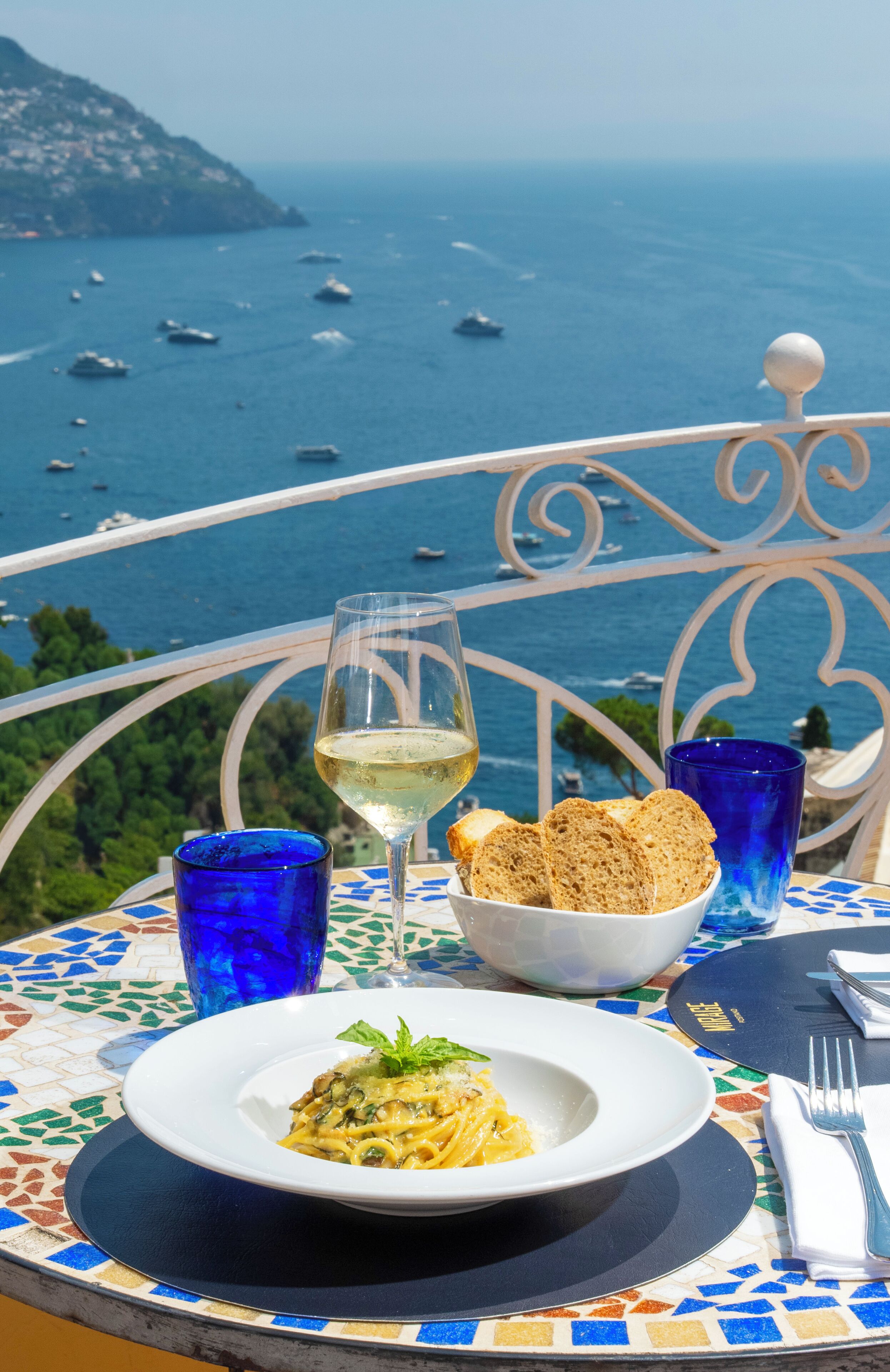 Una mesa con un plato de pasta, una copa de vino blanco y un bol de pan, frente a una vista panorámica del mar y barcos.