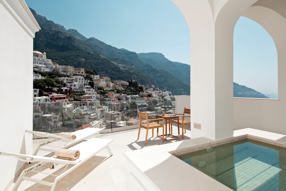 A pool area with a view of a hillside town, lounge chairs, and a small table.