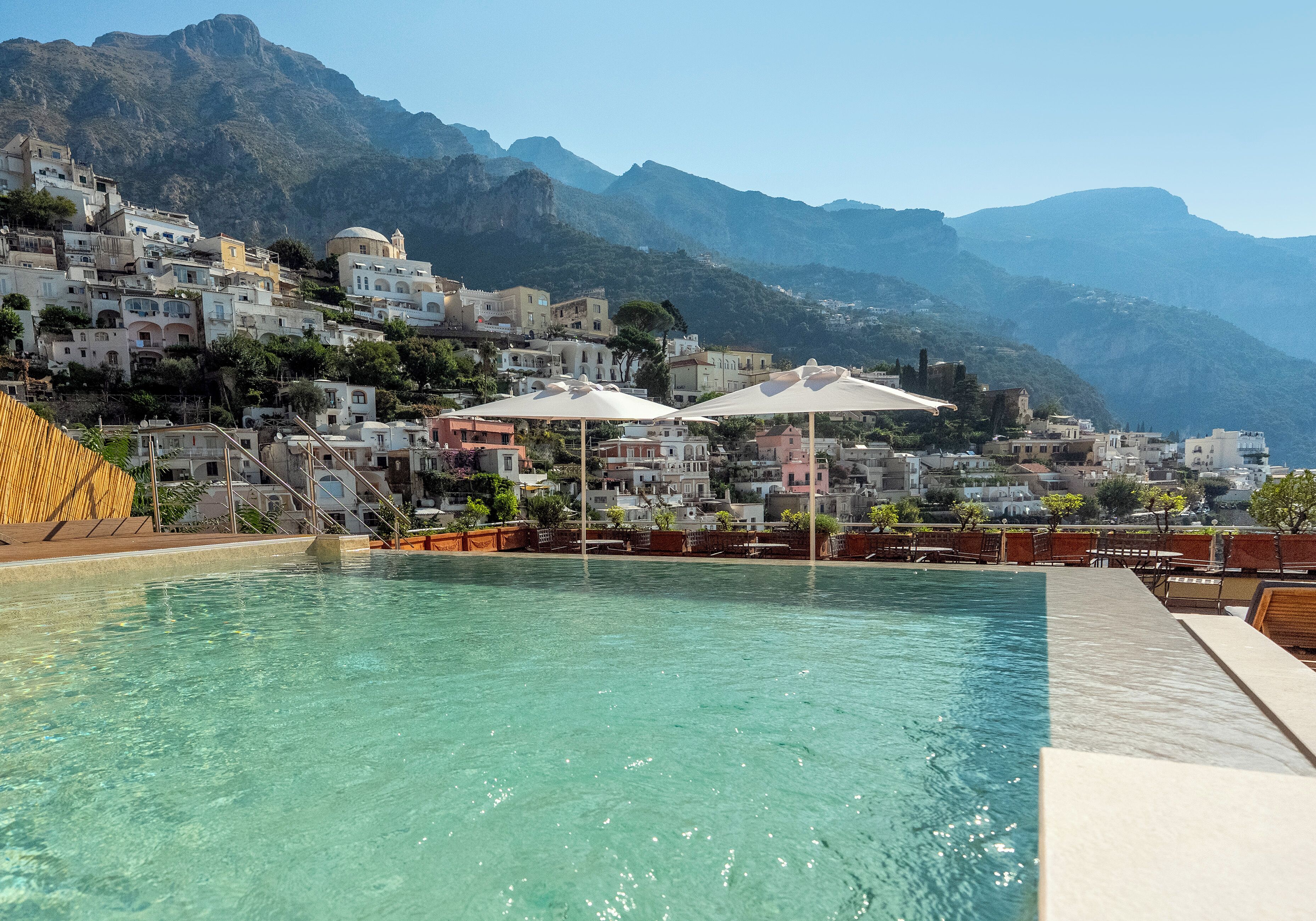 A swimming pool with clear water, surrounded by buildings and mountains in the background.