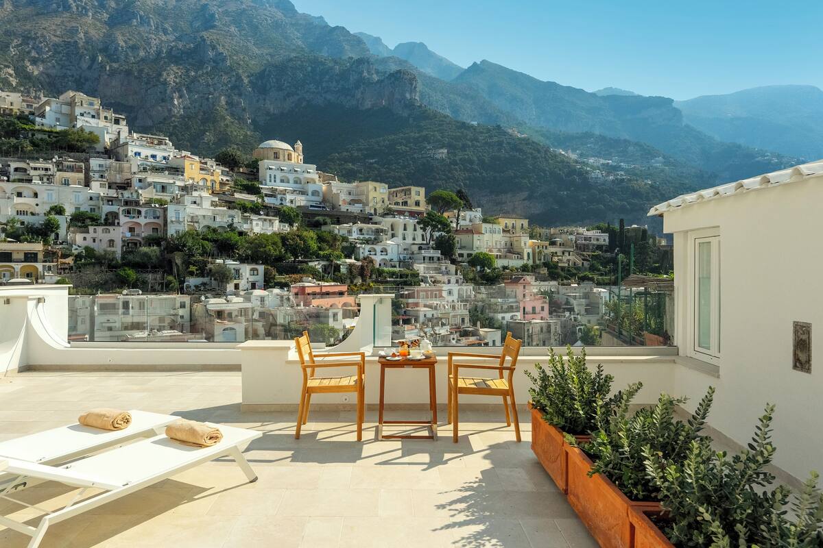 A rooftop terrace with a table and chairs, overlooking a mountainous town with terraced houses.