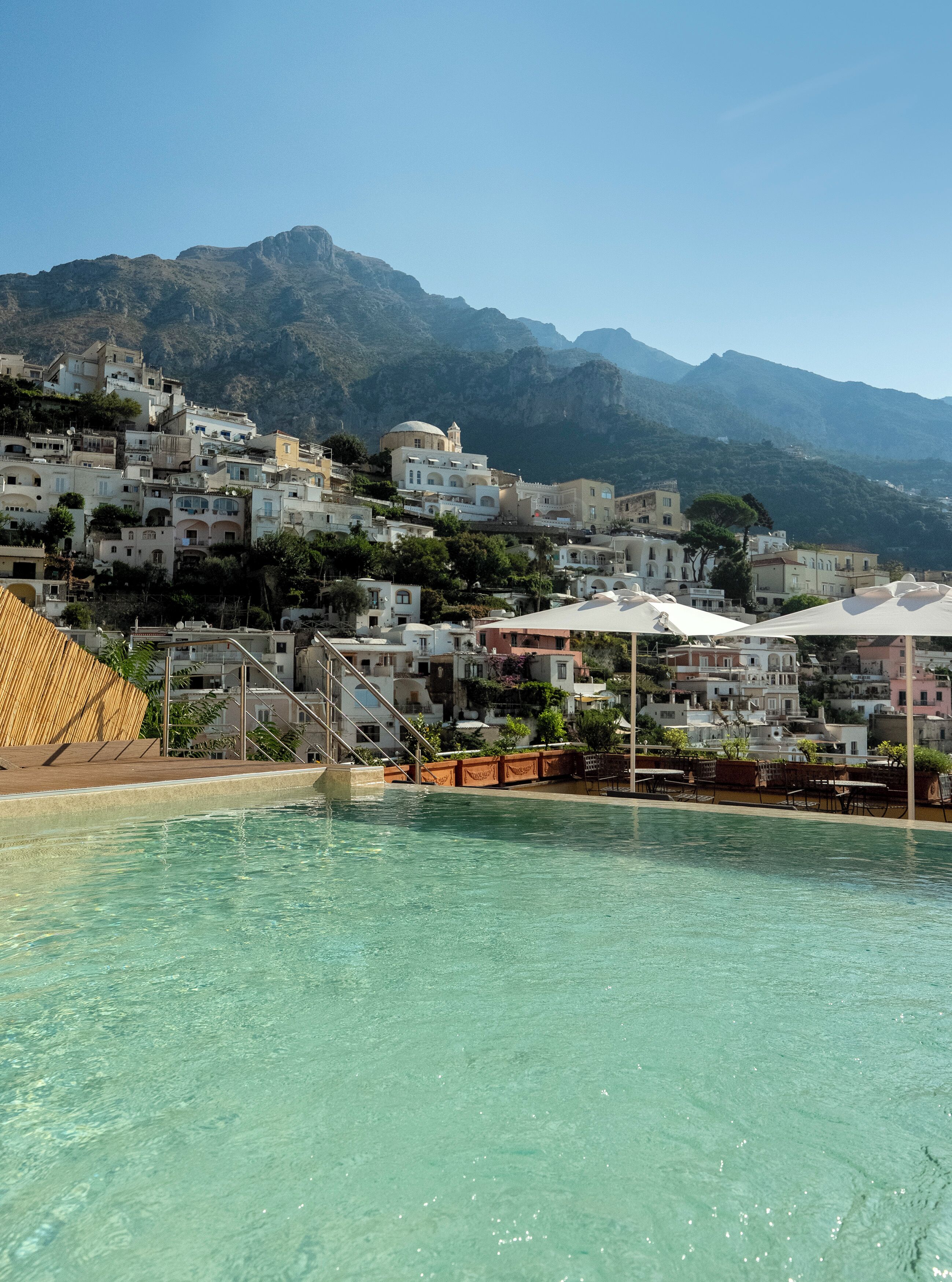 A pool area with clear water, surrounded by buildings with Mediterranean architecture, and mountains in the background.