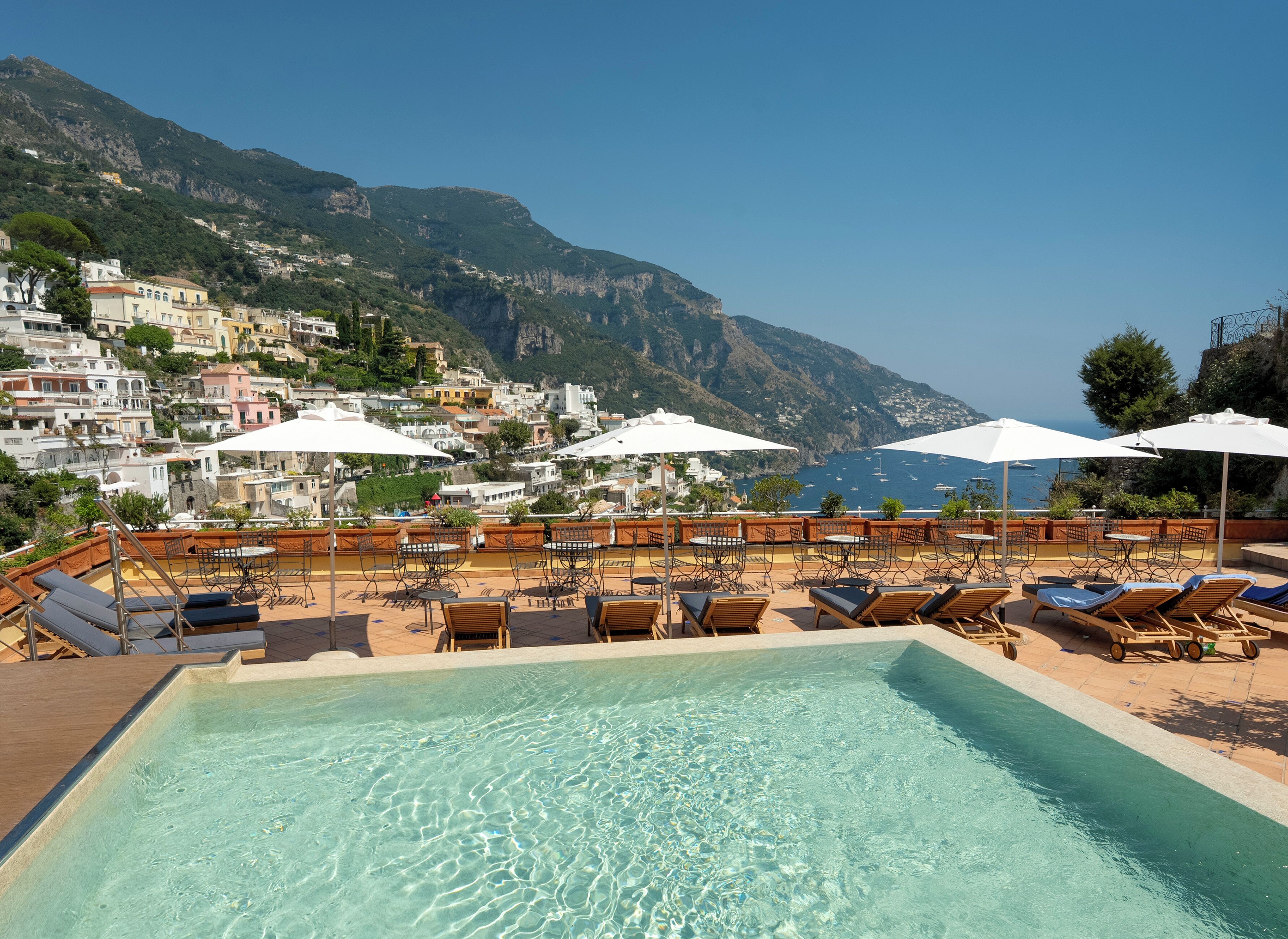 A poolside area with lounge chairs and umbrellas, overlooking a coastal town and mountains.