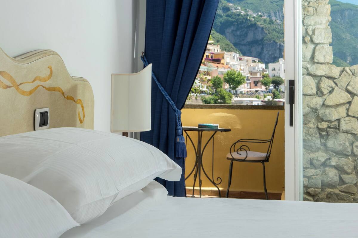 A hotel room with a bed, a nightstand, a lamp, a chair, and a balcony with a view of a hillside town.