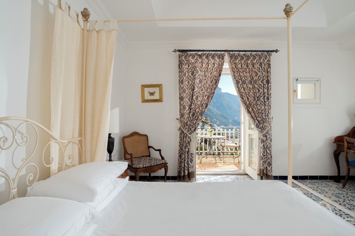 A bedroom with a four-poster bed, a chair, and a view of a balcony with a mountain in the background.