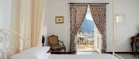 Quarto com cama de dossel, uma cadeira e vista para uma varanda com uma montanha ao fundo.