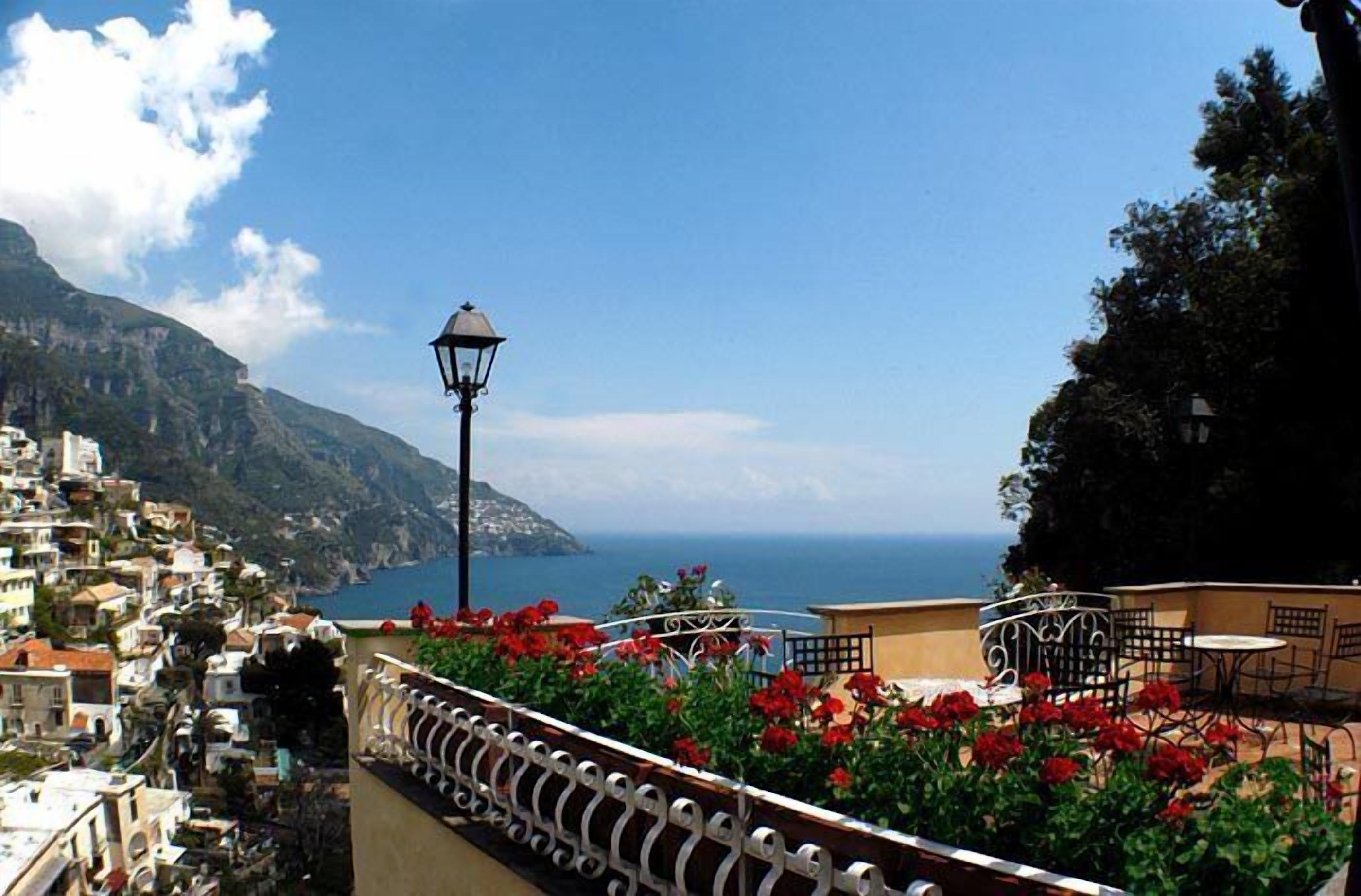 A coastal town with a balcony overlooking the sea, featuring a street lamp and red flowers.