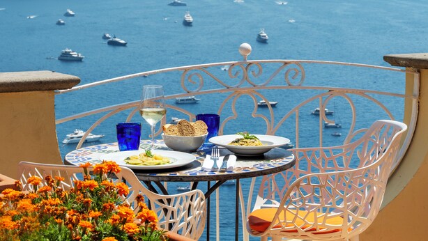 A table with plates of food, a glass of wine, and blue glasses on a balcony overlooking the sea with boats. - Hotel Posa Posa (Positano)