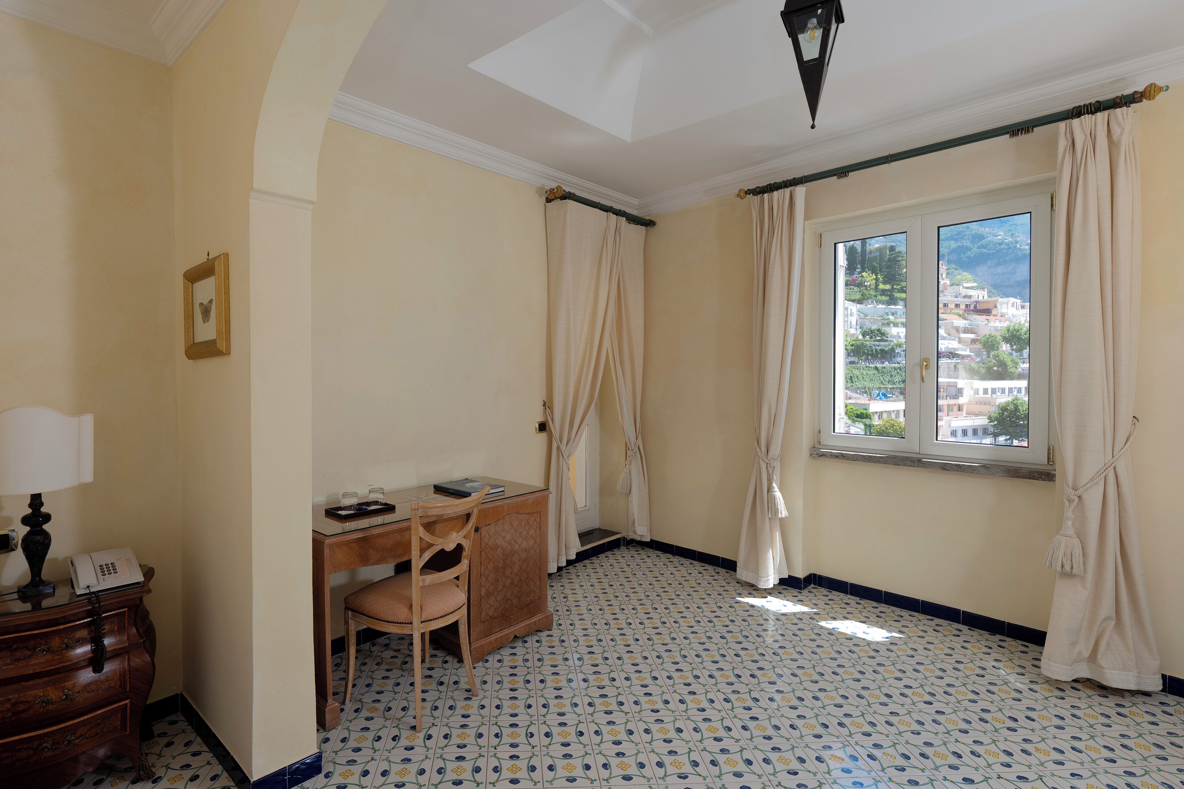 Una habitación con escritorio, silla y teléfono sobre una cómoda. Hay una ventana con cortinas y vistas a edificios y montañas.