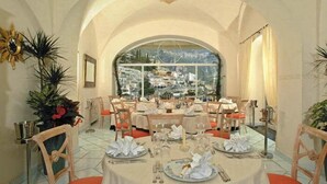 A dining area with tables set for a meal, a large arched window, and a view of a town. - Hotel Posa Posa (Positano)