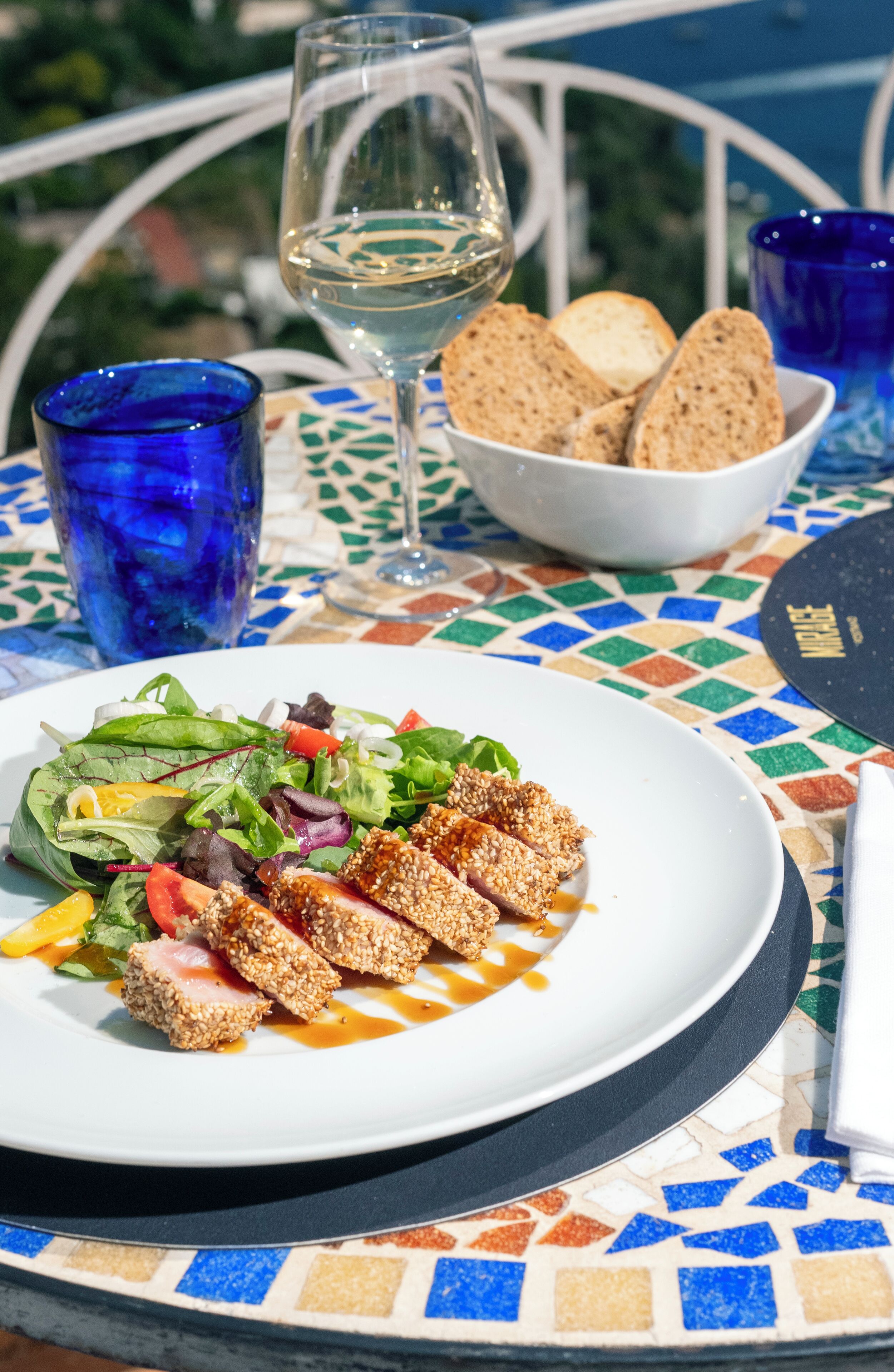 Una mesa con un plato de comida, una copa de vino blanco y un bol de pan.