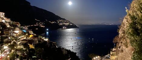A coastal town at night with a prominent moon in the sky, buildings along the coastline, and boats on the water.