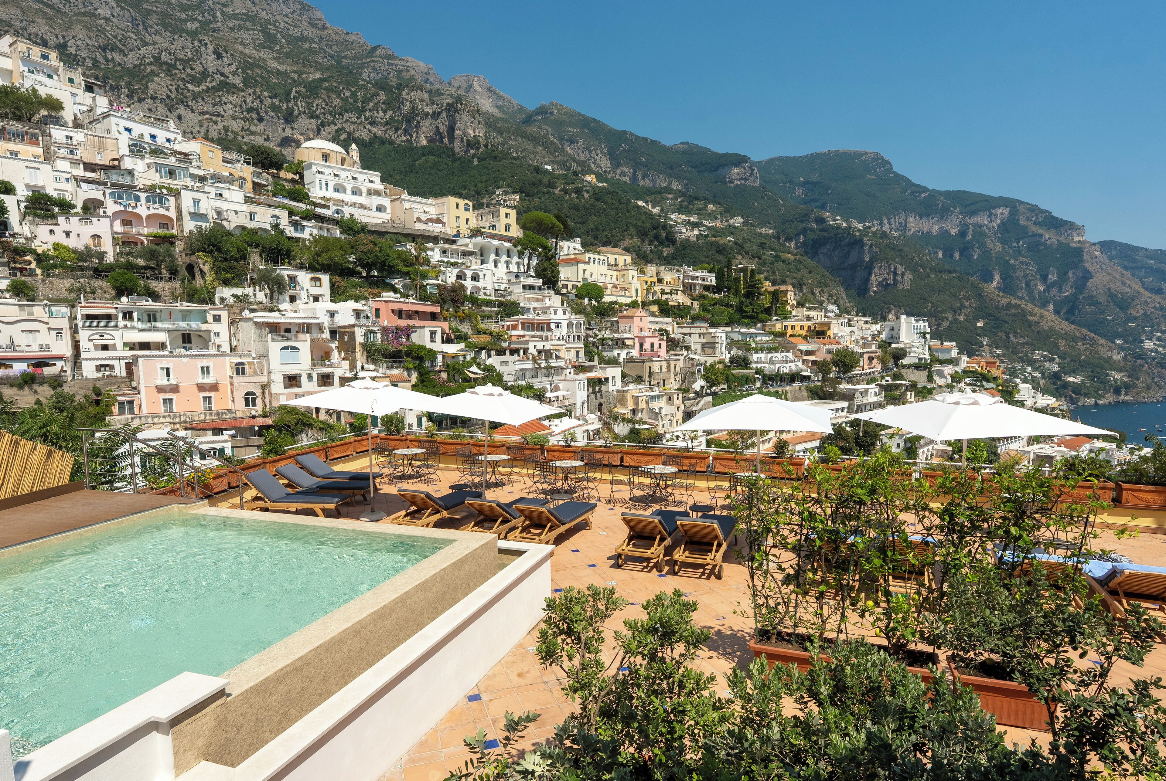 Un espace piscine aménagé sur le toit, avec des chaises longues et des parasols, offrant une vue sur une ville perchée sur une colline, avec ses bâtiments et un paysage montagneux en arrière-plan.