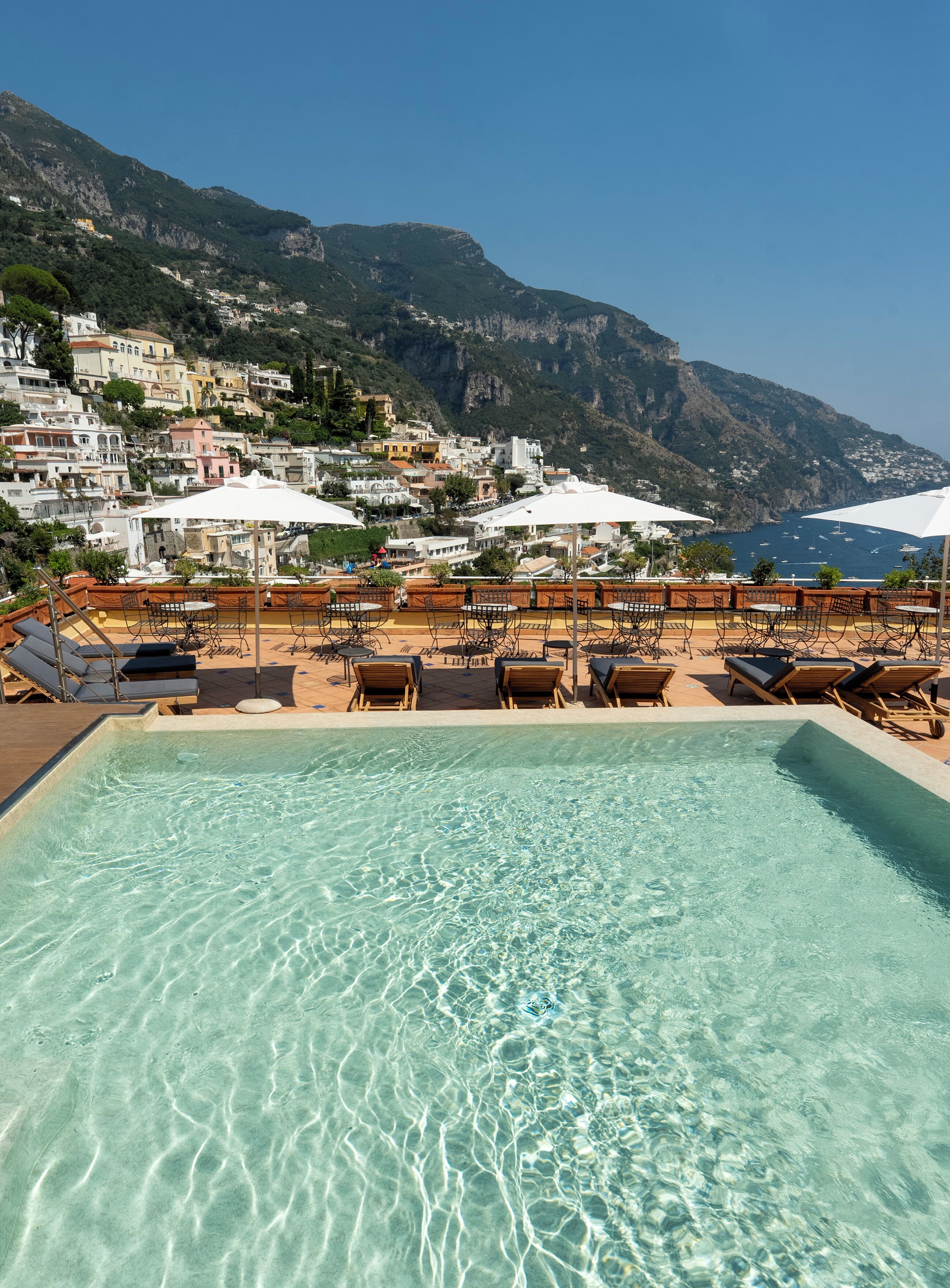 A pool area with clear water, lounge chairs, and umbrellas, overlooking a hillside town with buildings and a view of the sea.